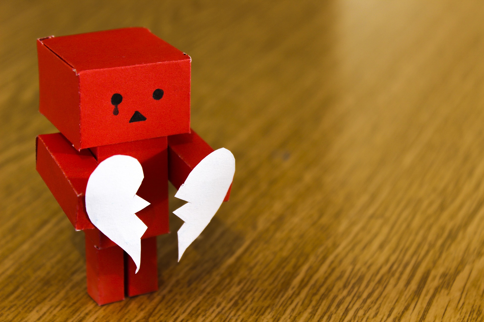 A sad little red paper robot holds a broken white paper heart